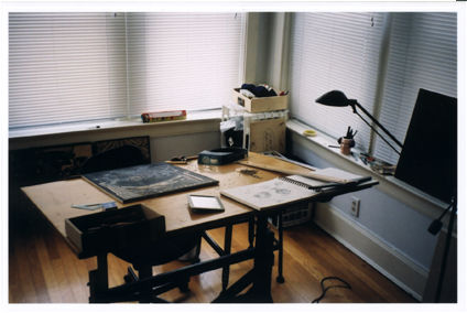 A desk sits infront of drawn mini-blind. Various art related instruments lie strewn about.
